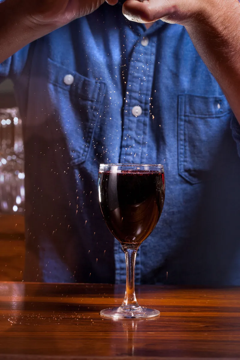Hands in a denim work shirt crumble garnish over a dark red drink, amber particles frozen mid-fall on a bar top.