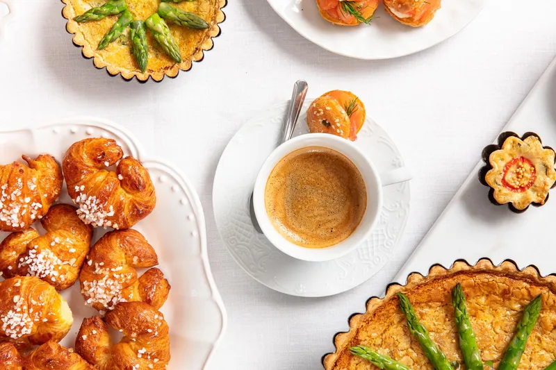 Espresso at the center of a family style breakfast spread with mini croissants, quiche, and smoked salmon bites