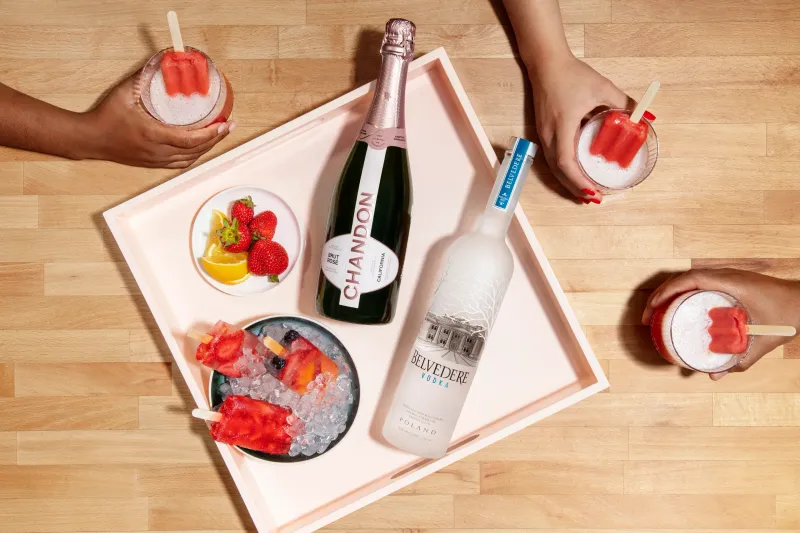 Top-down Chandon Brut Rosé and Belvedere Vodka bottles on a blush tray with hands holding frozen fruit cocktails.