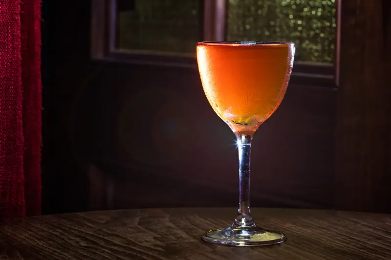 Amber stirred cocktail in a Nick and Nora glass, backlit beside the Dead Rabbit red curtain