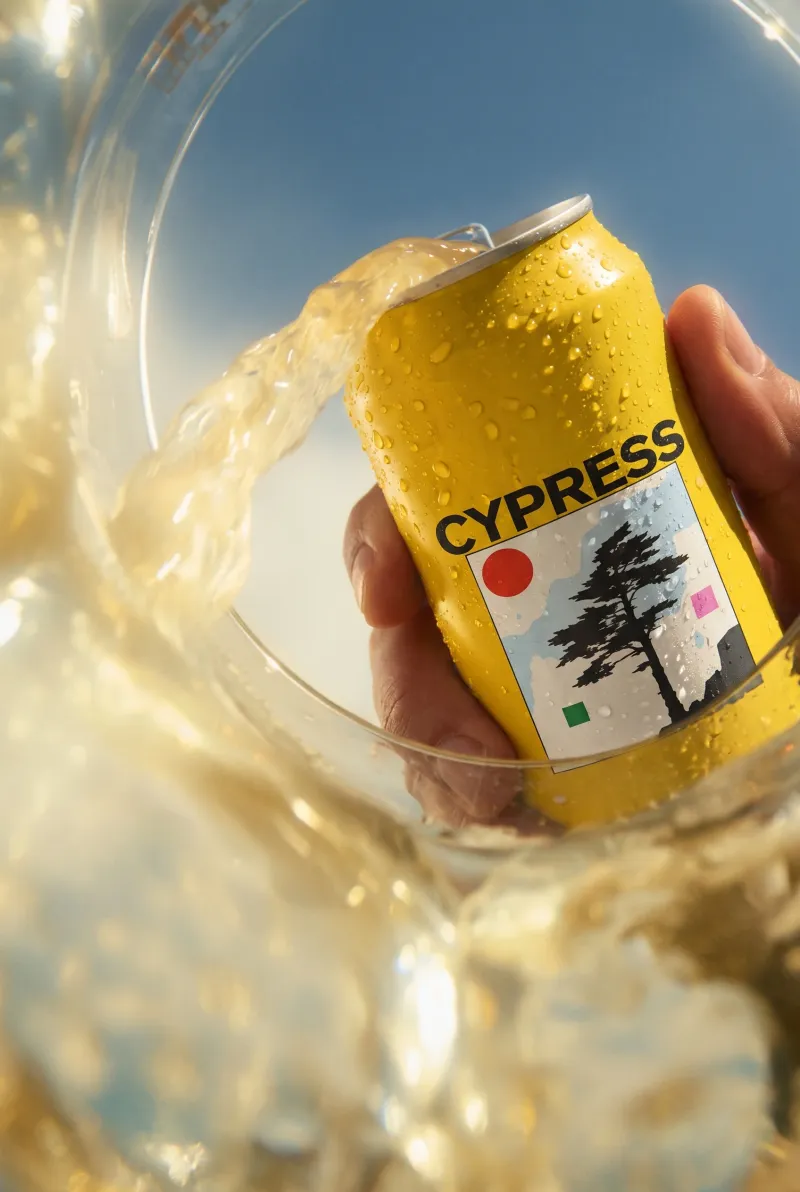 Cypress lager can held above a glass as beer splashes in under a blue sky, commercial beer photography