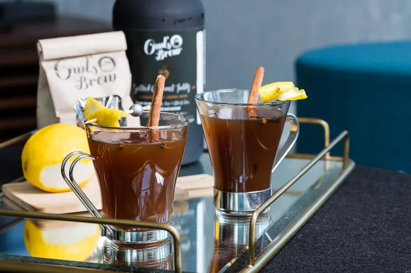 Two hot toddies with cinnamon sticks, cloves, and lemon twists on a mirrored tray beside an Owl's Brew bottle